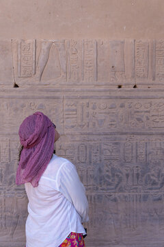Woman Solo traveler wearing the Egyptian turban or Imma, observing detailed carvings on a historical Philae temple wall in Aswan, immersed in egypt's rich cultural heritage and art