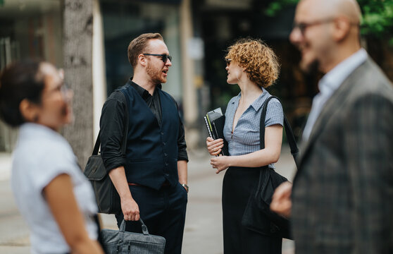 A group of business colleagues chat casually on a city sidewalk, holding bags and folders. They engage in friendly conversation between appointments in an urban workday setting.