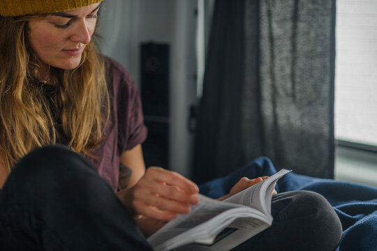 Woman wearing a yellow beanie reads a book in her van