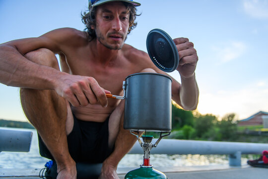 Shirtless man cooking food on a camp stove by a lake