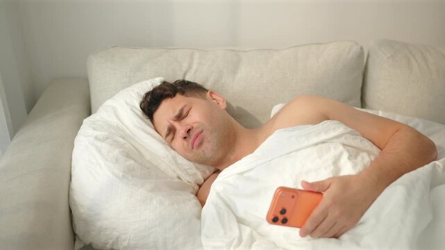 A shirtless man lying on a sofa bed grimaces at his orange phone, then squeezes his eyes shut and tucks it under the white duvet. Lazy morning in a bright bedroom with sheer curtains and soft light.