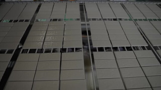 Grey pressed tiles sit in neat rows on a steel drying rack inside a busy ceramic works