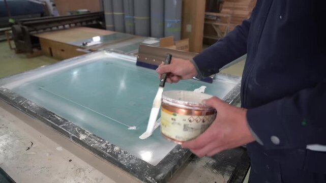 Worker spreading white ink along the edge of a large silkscreen frame with a spatula before a print run