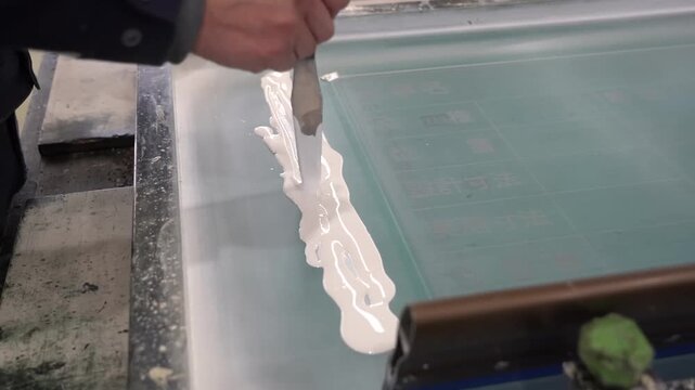 Craftsman laying a thick line of white ink onto a silkscreen mesh with a spatula ready to be pulled across