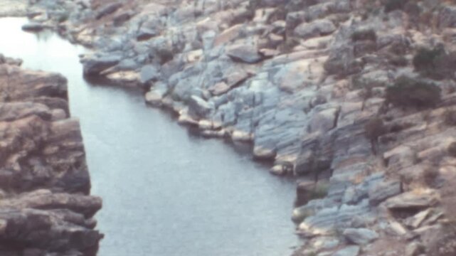 Narrow river flows through a steep rocky gorge in Portugal. Calm dark water winds between layered grey rocks and sparse dry scrub vegetation in this rugged arid natural landscape archival footage.