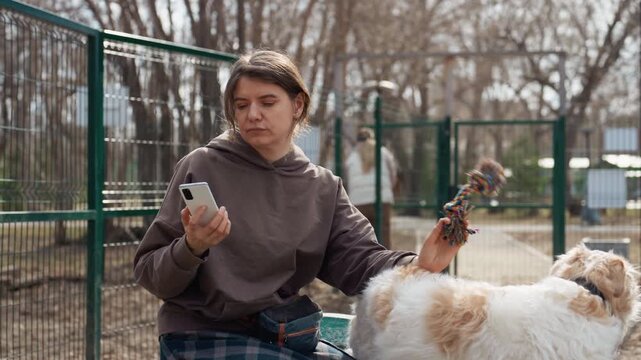 Woman sits outdoors holding smartphone in one hand and interacting with her dog who is playing or resting beside her. Woman wears casual clothing suitable for outdoor setting likely at park