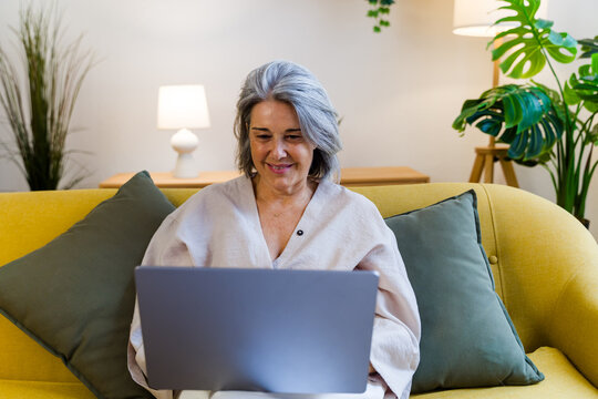 Senior white woman using laptop on yellow sofa in modern living room