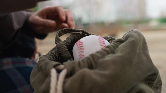 Person places baseball and colorful fidget toys into olive green drawstring bag then secures closure. focus remains on hands interacting with fabric textures in soft natural light