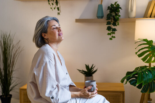 White woman sitting indoors holding mug and thinking