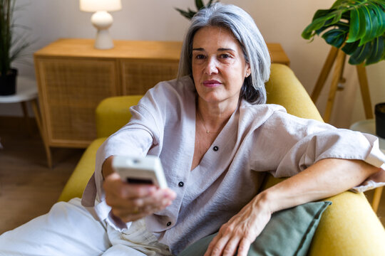 Mature white woman holding remote control on yellow couch indoors