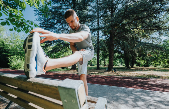 Young man performs a leg stretch on a park bench as part of his fitness routine. He warms up outdoors before running, demonstrating a focused pre-workout stretch.