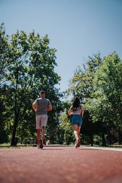 A man and woman jog together along a sunlit park path. The couple runs side by side surrounded by trees and blue sky.
