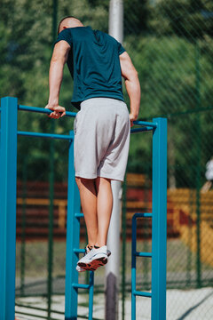 A young man performs a dip exercise on blue parallel bars at an outdoor fitness area. The athlete trains calisthenics on a pull-up and dip station in a sunny park.