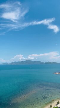 Vertical high-angle view of the beautiful blue ocean bay in Nha Trang, Vietnam, featuring distant green islands and a sunny sky with wispy white clouds.