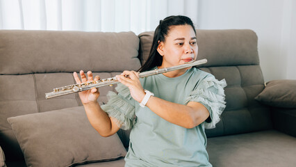 A female musician plays a silver flute in her home, standing near a gray sofa. Her green shirt and the warm living room decor make for a cozy yet vibrant musical atmosphere. © sorapop
