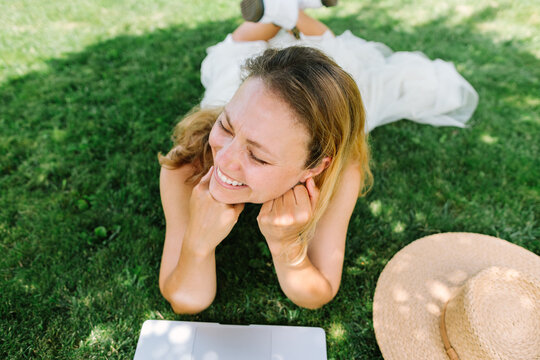 Smiling woman works on a laptop while lying on the grass outdoors