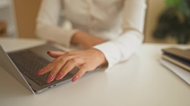 Young woman in white blouse typing on laptop with manicured hands at glossy desk surrounded by notebook papers folders and plant in building; concentration productivity.