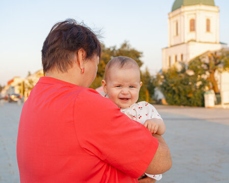 woman carrying baby over shoulder in calm outdoor urban scene with church in background, everyday family portraits and parent-child relationships visual resource.