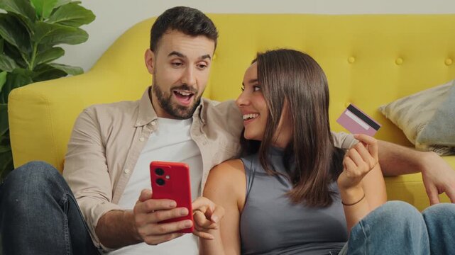 A cheerful pair lounges on a bright yellow sofa as the man uses a red phone and the woman holds a card, suggesting financial or shopping themes with a warm home vibe and natural light.