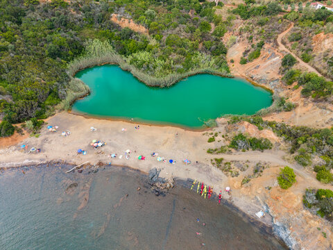 Aerial view of Laghetto di Terra Nera, a turquoise lake by a sandy beach with colorful umbrellas and kayaks on the shore in Porto Azzurro, Tuscany, Italy.