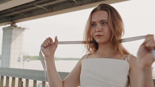 Woman stands on bridge holding and measuring herself with tape measure against her body. background features outdoor riverside setting with soft daylight lighting and architectural elements