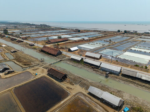 Aerial view of Salt Ponds featuring rectangular evaporation basins, traditional wooden storage sheds with tiled roofs, and a narrow canal in Kedung, Central Java, Indonesia.