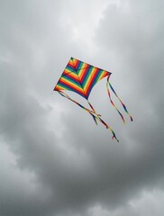 Fototapeta premium Bright rainbow striped kite flies high in moody grey overcast sky. Colorful toy with long tails floats against dark clouds. Wind carries diamond shaped kite during windy autumn summer day. Simple