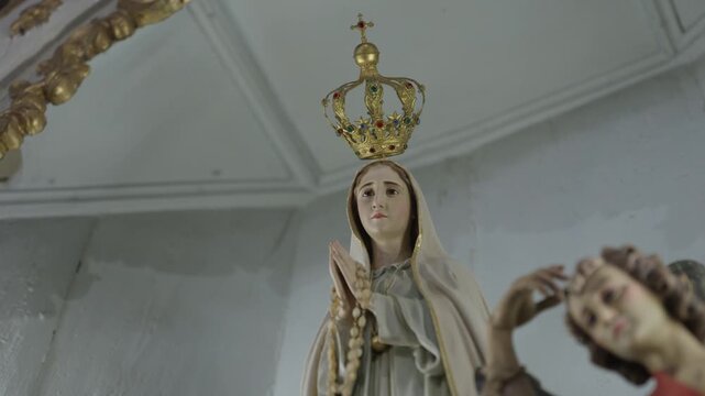 A statue of Our Lady of Fatima wearing a gold jeweled crown and holding a rosary, seen from below in a white chapel.