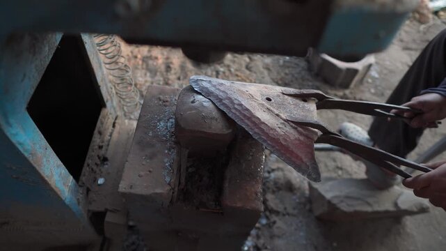 Close-up of a blacksmith using forging tongs to hold heated metal on an anvil beneath a mechanical power hammer during a metalworking and shaping process.