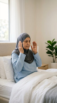 Senior Muslim woman in a grey hijab performing morning prayer or dua while sitting on a bed in a bright and minimalist bedroom interior background.