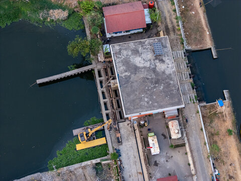 Aerial view of a water management structure with a sluice gate, a yellow excavator parked on the bank, nearby buildings, and water channels in Semarang, Central Java, Indonesia.