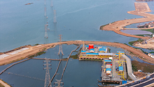 Aerial view of an industrial facility with storage tanks and pipelines situated along the coastline, featuring power transmission towers extending into the water in Semarang, Central Java, Indonesia.