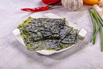 White ceramic plate holding dark green nori seaweed sheets with red chili pepper, garlic bulb, and green onions in background, food ingredient © Andrei Starostin