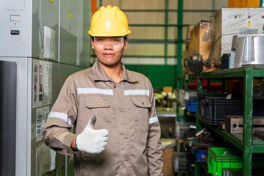 Happy warehouse operator giving thumbs up sign surrounded by storage racks and equipment, smiling asian factory worker showing thumbs up gesture in manufacturing warehouse plant