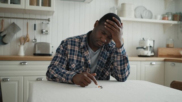 Depression and illness, african man looking at different pulls on kitchen table. Medium portrait of sad black male person, medications and side effect, domestic chemotherapy for cancer, curing illness