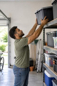 Young man placing storage bin with holiday decorations on high shelf in organized home garage, january cleanup