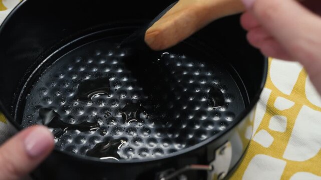 Close-up of a hand greasing a round black springform baking pan with oil using a wooden-handled pastry brush, homemade baking step. High quality 4k footage