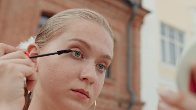Young woman receives makeup application outdoors focusing on eye and face details. hand applies mascara near her ear while another holds compact mirror with red accents. background features