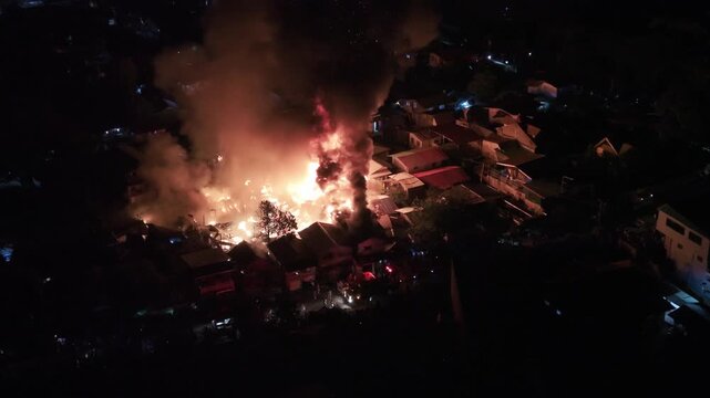 Philippines, Palawan, Puerto Princesa 10.05.2026: Close aerial night view of houses engulfed in flames in Barangay Masipag, with thick smoke, glowing roofs and emergency lights on the street below.