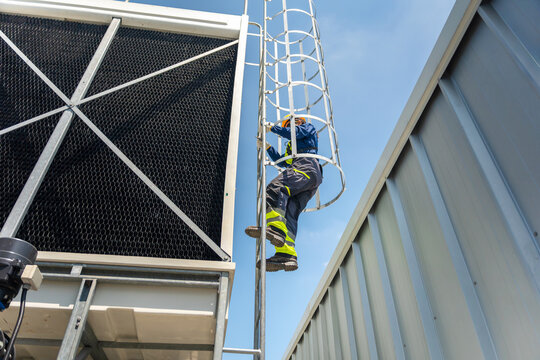 Industrial worker climbing safety cage ladder to rooftop cooling tower for maintenance, maintenance engineer climbing safety access ladder near large HVAC air conditioning unit