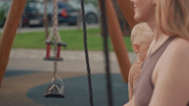 Young boy stands near playground swing interacting with adult woman. scene takes place outdoors in daylight at park or backyard setting featuring swings and soft focus background elements