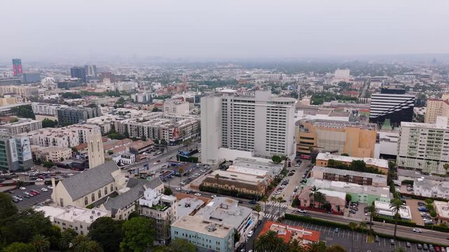 Hollywood district in Los Angeles from a drone. A gothic church with a bell tower, a white high-rise and palm trees along the boulevard. Hazy overcast sky over the city sprawl and distant skyline.