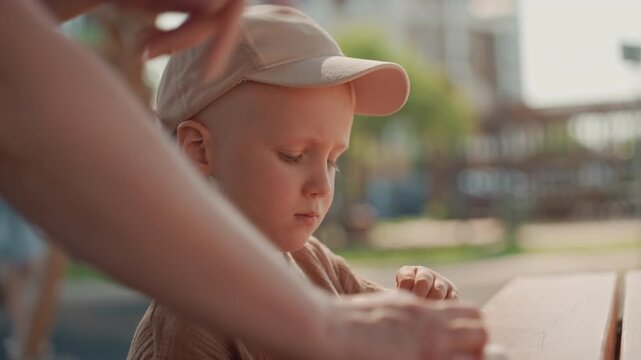 Young boy wearing beige cap and brown shirt examines or manipulates object on wooden table outdoors. Another boy arm is visible suggesting interaction with child in this casual setting