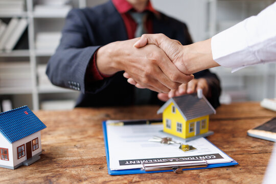 Business partners shaking hands across a wooden desk with house models, keys, and a contract, representing a finalized agreement for property investment and home ownership
