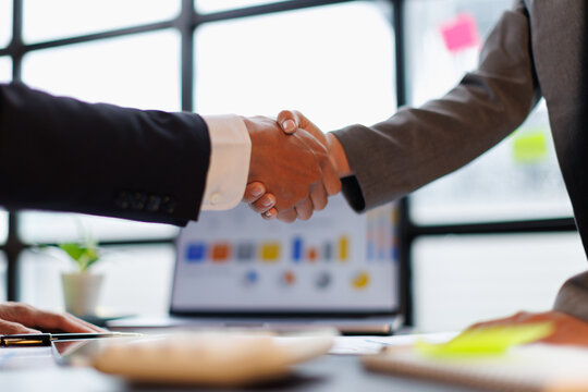Business professionals shaking hands over a desk, sealing a partnership and agreement amid charts and laptops, symbolizing teamwork, trust, and corporate success