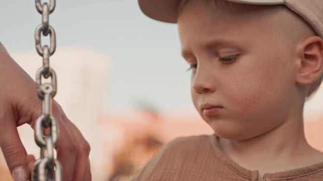 Young boy holds onto metal swing chain with adult hand nearby suggesting gentle interaction in soft daylight. scene focuses on close up details of their hands and part of child face