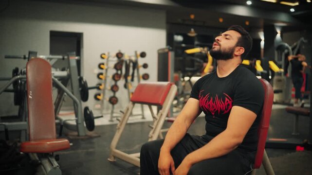 High quality interior video shot capturing a bearded man sitting upright on a gym bench, closing his eyes to recover from physical exertion and muscle exhaustion.