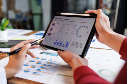 Business professionals reviewing financial and marketing reports on a tablet and printed documents, pointing with a pen at charts and graphs to assess growth and performance