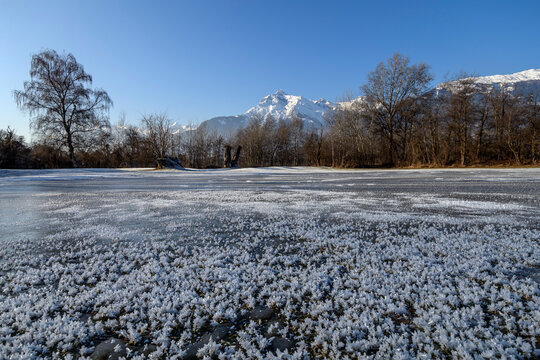 Close-up details of delicate frost crystals on a frozen lake surface with snow-covered alpine mountains