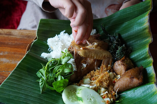 Hand gently arranges rice, greens, and meats on a banana leaf. Fruit, herbs, and savory meats rest atop the leaf. Warm tones highlight the rustic dining experience